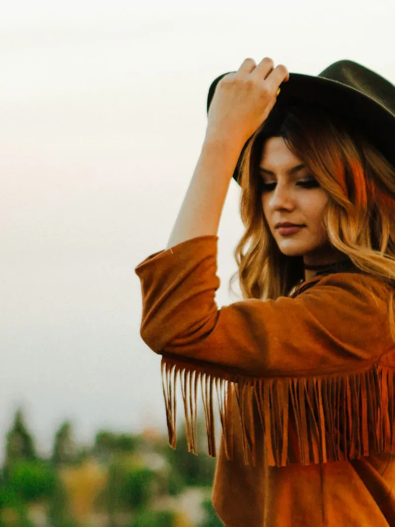 Tan fringe suede jacket worn with a black felt hat at golden hour
